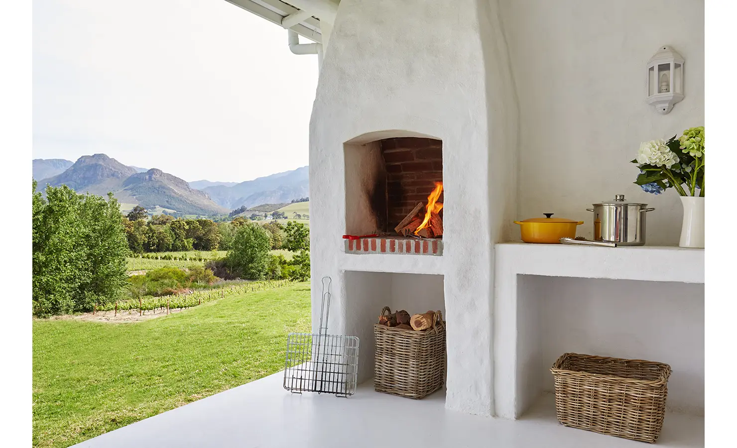 Perfect Hideaways: An outdoor braai (barbecue) area with a lit brick fireplace, wicker baskets, and a scenic view of vineyards and mountains. La Cotte Forest Cottages, Franschhoek.