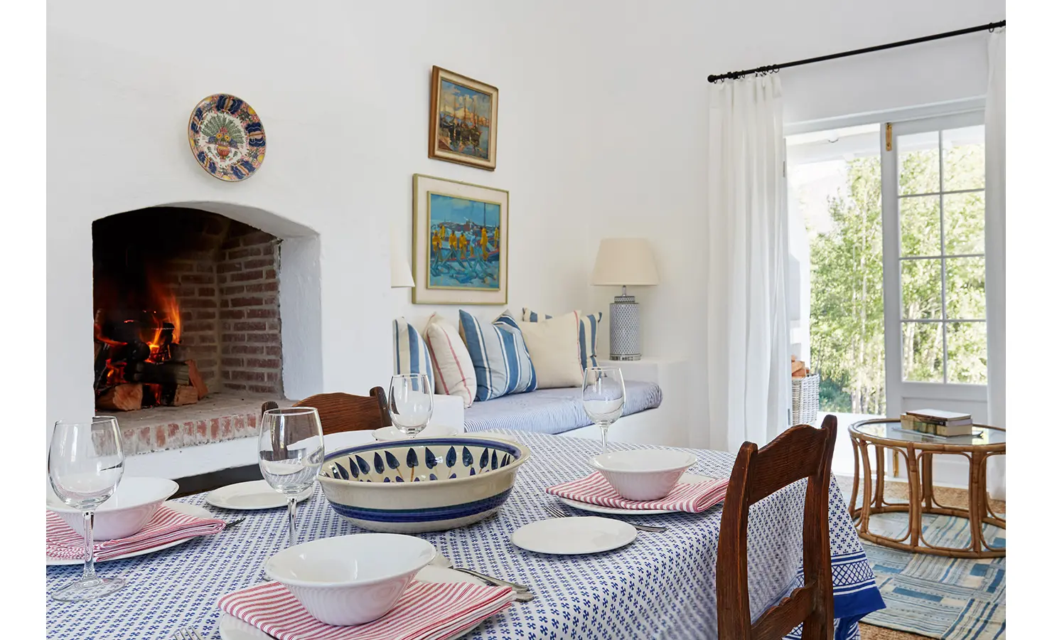 Perfect Hideaways: A dining area with a table set for four, featuring blue-and-white accents, and a lit fireplace, with a view of a cosy lounge in the background. La Cotte Forest Cottages, Franschhoek.