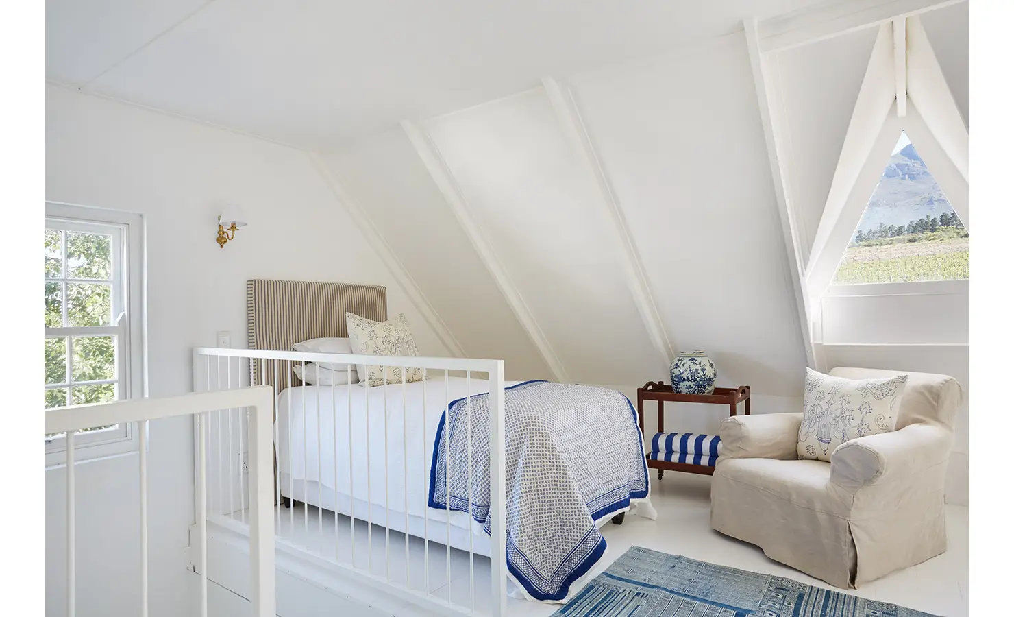Perfect Hideaways: An attic-style bedroom with a pitched ceiling, white bedding with blue accents, and a comfortable armchair by a triangular window showcasing mountain views. La Cotte Forest Cottages, Franschhoek.