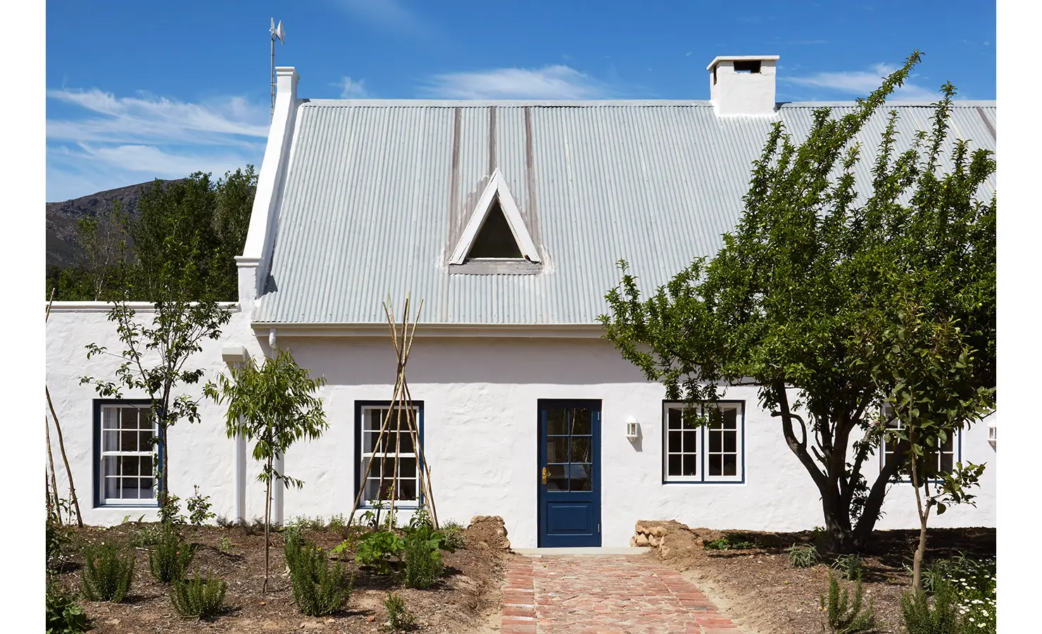 Perfect Hideaways: An exterior view of a charming white cottage with a tin roof, blue windows, and a lush garden with neatly planted greenery. La Cotte Forest Cottages, Franschhoek.