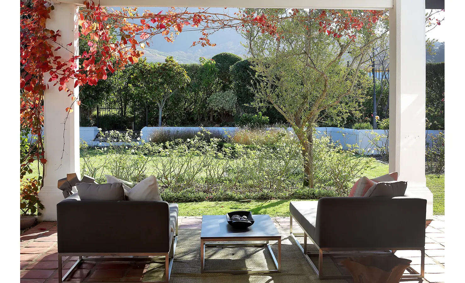 Perfect Hideaways: A peaceful garden veranda framed by vibrant autumnal vines, offering a pair of modern loungers with plush cushions and serene views of the lush landscape. Buitenzorg Estate, Constantia.