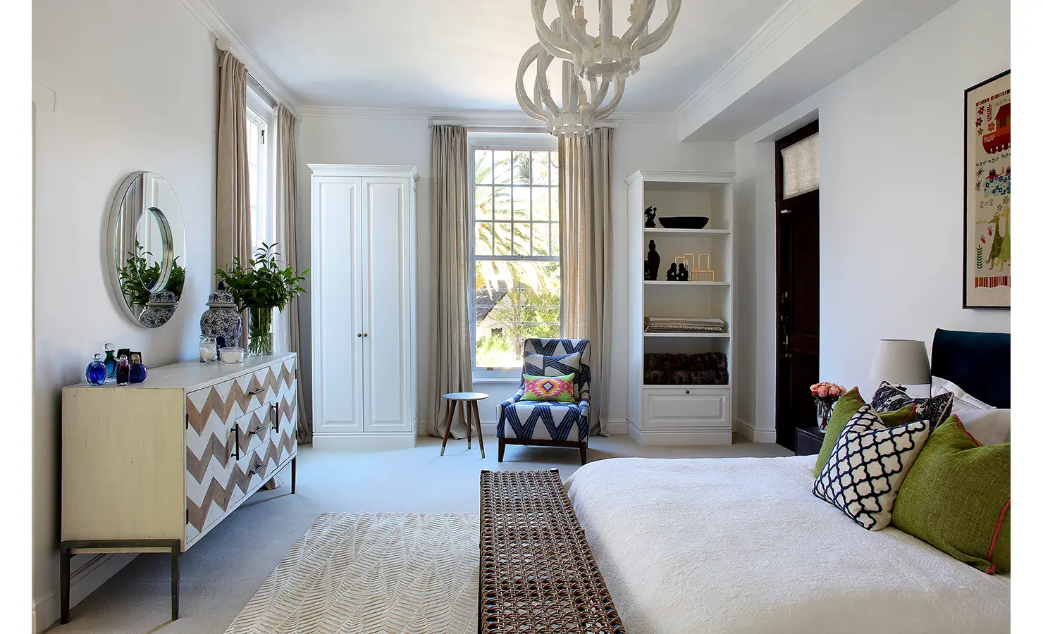 Perfect Hideaways: A tranquil bedroom in soft neutral tones, featuring an elegant wooden dresser, a chevron-patterned sideboard, and a cosy reading nook with a patterned armchair. Sunlight streams through tall windows, enhancing the serene feel. Buitenzorg Estate, Constantia.