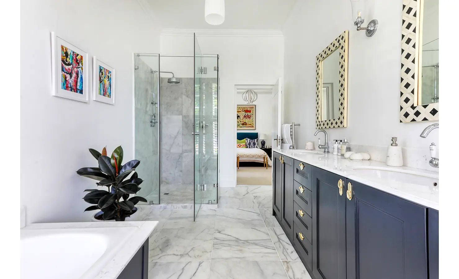 Perfect Hideaways: A light-filled bathroom with sleek marble flooring, a glass-enclosed shower, and a deep double vanity with navy cabinetry and brass accents. A view through the doorway reveals a luxurious bedroom beyond. Buitenzorg Estate, Constantia.