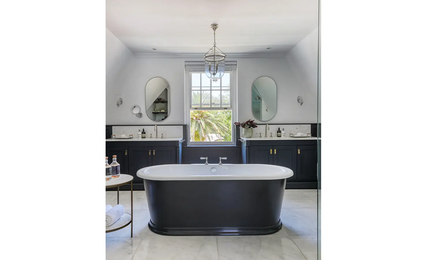 Perfect Hideaways: A sophisticated bathroom with a deep black freestanding tub, dual vanities, and a vintage-style pendant light. A large window frames lush greenery, blending elegance with nature. Buitenzorg Estate, Constantia.