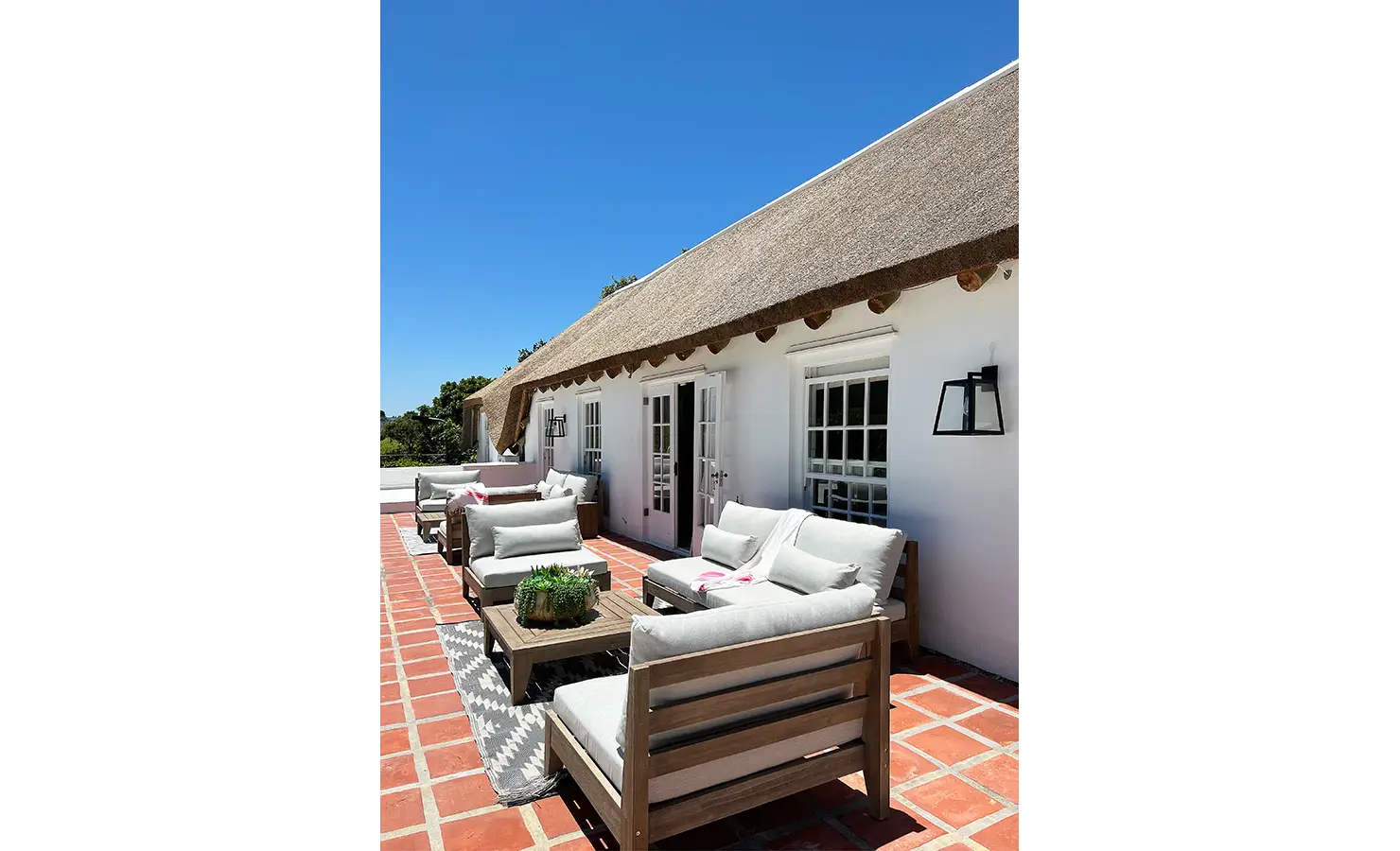Perfect Hideaways: A sun-drenched rooftop terrace with a thatched overhang, featuring plush outdoor seating in neutral tones. The terracotta-tiled flooring adds warmth against the brilliant blue sky. Buitenzorg Estate, Constantia.