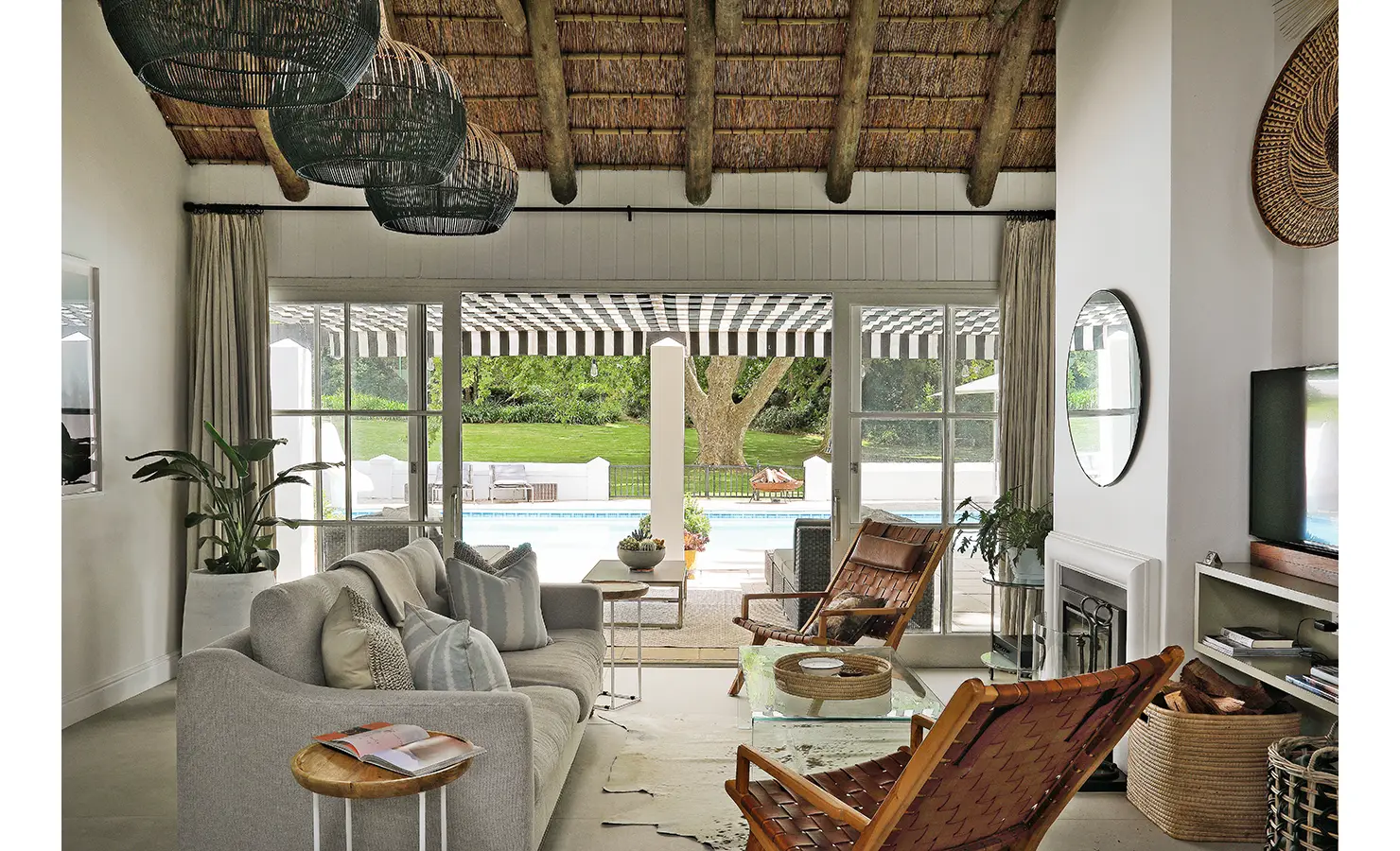 Perfect Hideaways: A beautifully designed living space with a thatched ceiling, woven pendant lights, and large glass doors opening to a poolside terrace with green lawns beyond. Buitenzorg Estate, Constantia.
