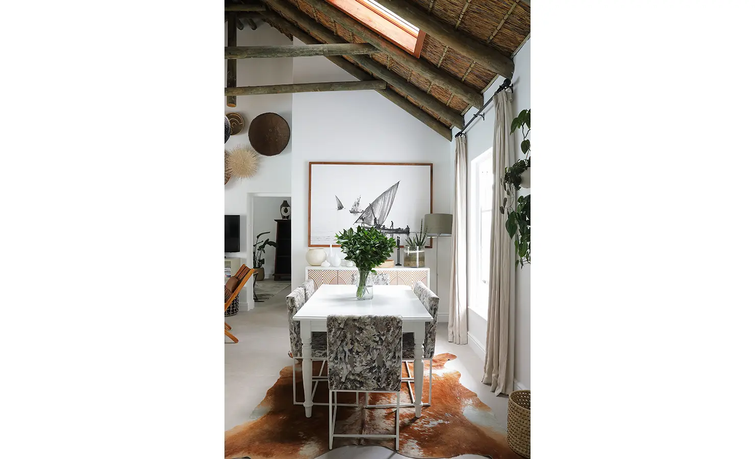 Perfect Hideaways: A chic dining area with a cowhide rug, botanical centrepiece, and a large framed artwork of traditional sailboats, set beneath a rustic thatched roof. Buitenzorg Estate, Constantia.