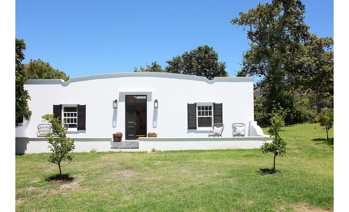 Perfect Hideaways: A charming white-washed Cape Dutch-style cottage with a gently curved gable, black shutters, and a lush green lawn. Two wicker chairs sit on the front stoep, creating an inviting outdoor space. Buitenzorg Estate, Constantia.