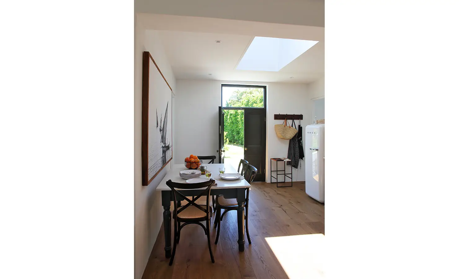 Perfect Hideaways: A bright and airy dining space featuring a rustic wooden table set for two, with a bowl of fresh oranges and green glasses. Natural light floods in through a skylight and an open black door leading to the lush garden. Buitenzorg Estate, Constantia.