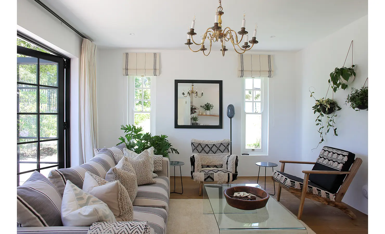 Perfect Hideaways: A beautifully curated living room with a mix of neutral tones, striped and textured cushions, and bohemian-style armchairs. A classic brass chandelier hangs from the ceiling, while large windows frame views of the surrounding greenery. Buitenzorg Estate, Constantia.