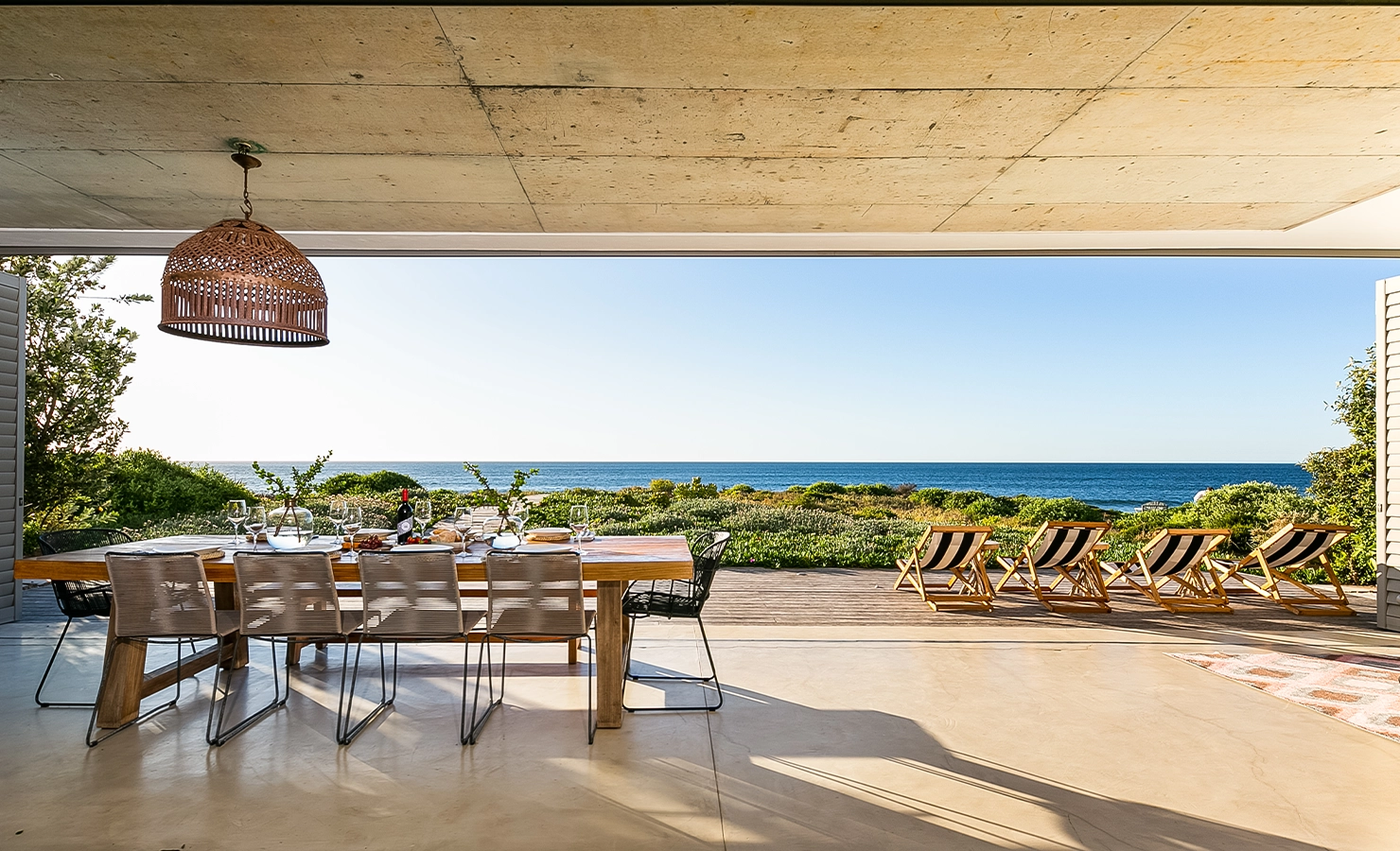 Perfect Hideaways: Indoor–outdoor dining space with a long wooden table set for lunch, woven pendant light, and striped deck chairs facing a wide sea view, Marrakesh House, Britannia Bay