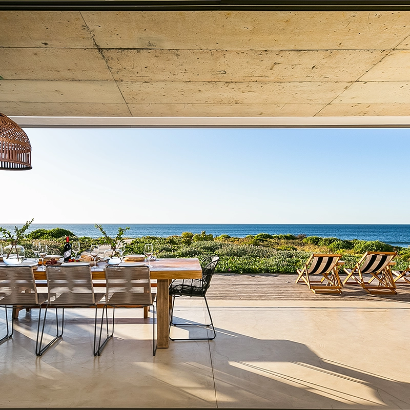 Perfect Hideaways: Indoor–outdoor dining space with a long wooden table set for lunch, woven pendant light, and striped deck chairs facing a wide sea view, Marrakesh House, Britannia Bay