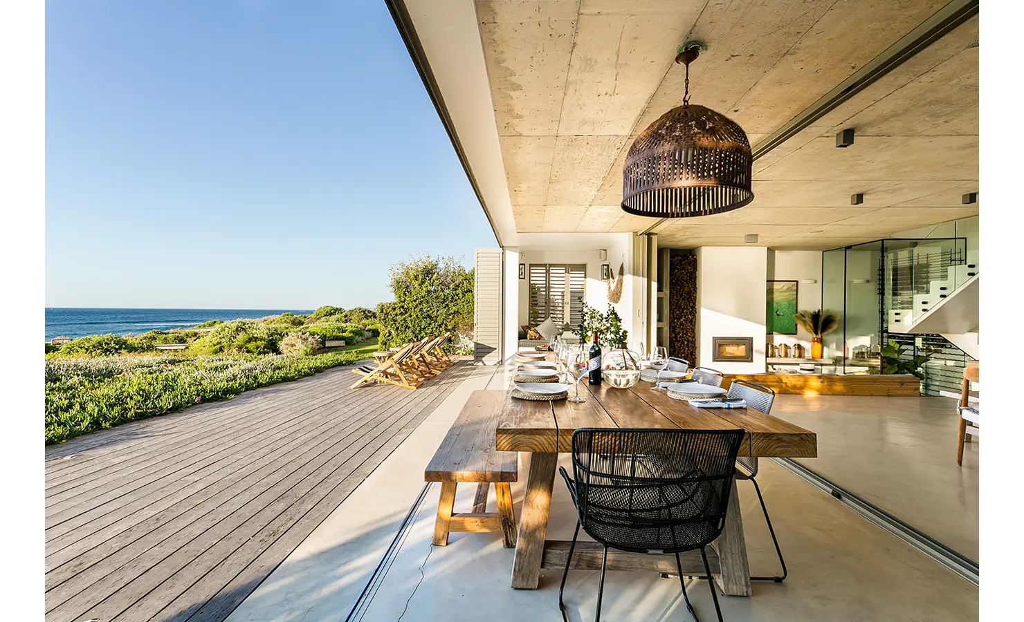 Perfect Hideaways: Ocean-facing indoor–outdoor dining area with a long wooden table, woven pendant and striped deck chairs on the timber deck, Marrakesh House, Britannia Bay