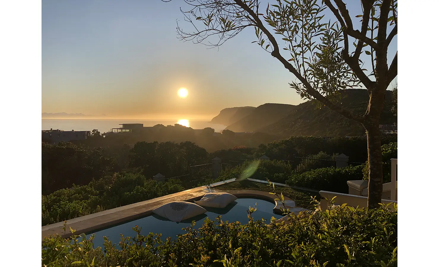 Perfect Hideaways: A serene view of the sun setting over the ocean, framed by lush greenery and a pool in the foreground, with distant cliffs and houses silhouetted against the golden sky. Beachwood On Robberg, Plettenberg Bay.