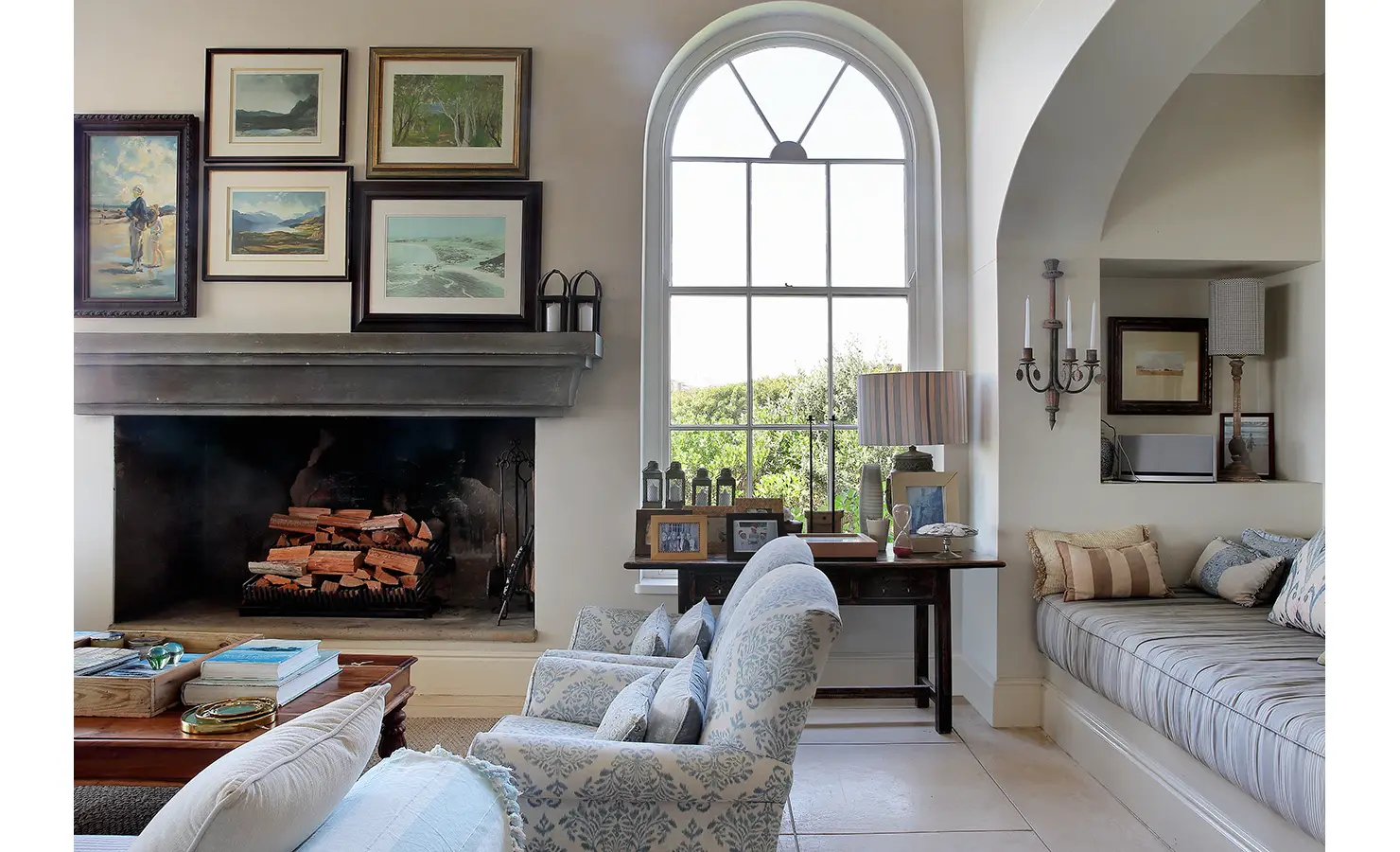 Perfect Hideaways: A warm and inviting sitting area with a stone fireplace, an arched window, and a mix of classic and coastal decor, including framed artworks. Beachwood On Robberg, Plettenberg Bay.