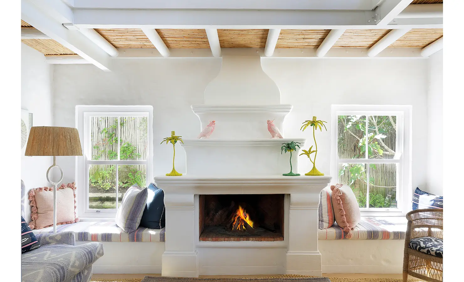 Perfect Hideaways: A striking white fireplace topped with playful beaded palm trees and parrots, flanked by window seats and pastel cushions in a bright, airy living space. Lalapanzi, Plettenberg Bay.