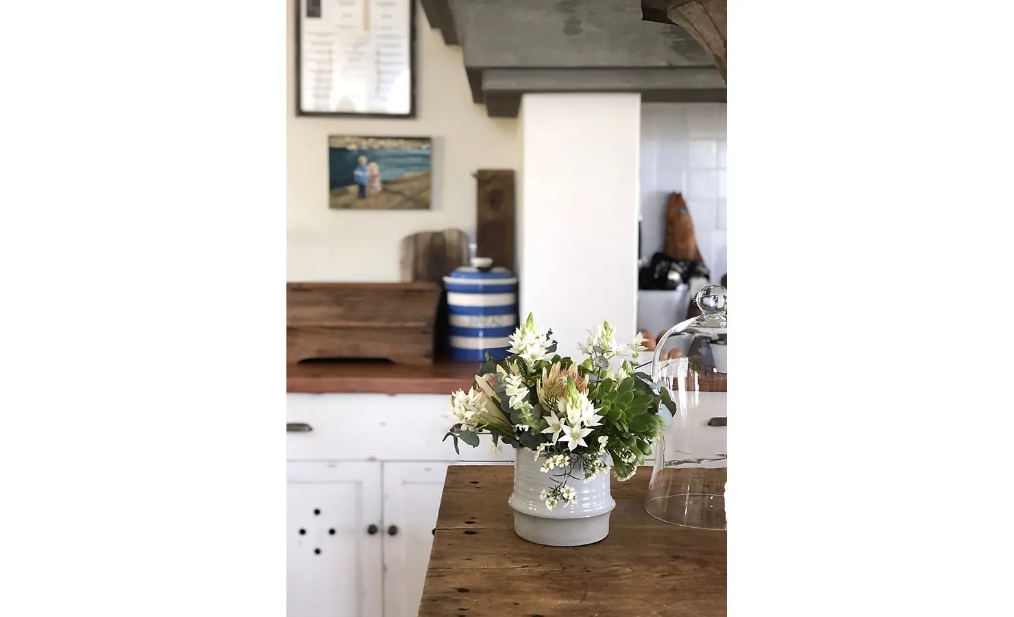 Perfect Hideaways: A close-up of a rustic kitchen countertop adorned with a ceramic vase holding fresh white flowers, complementing the wooden and vintage decor in the background. Beachwood On Robberg, Plettenberg Bay.