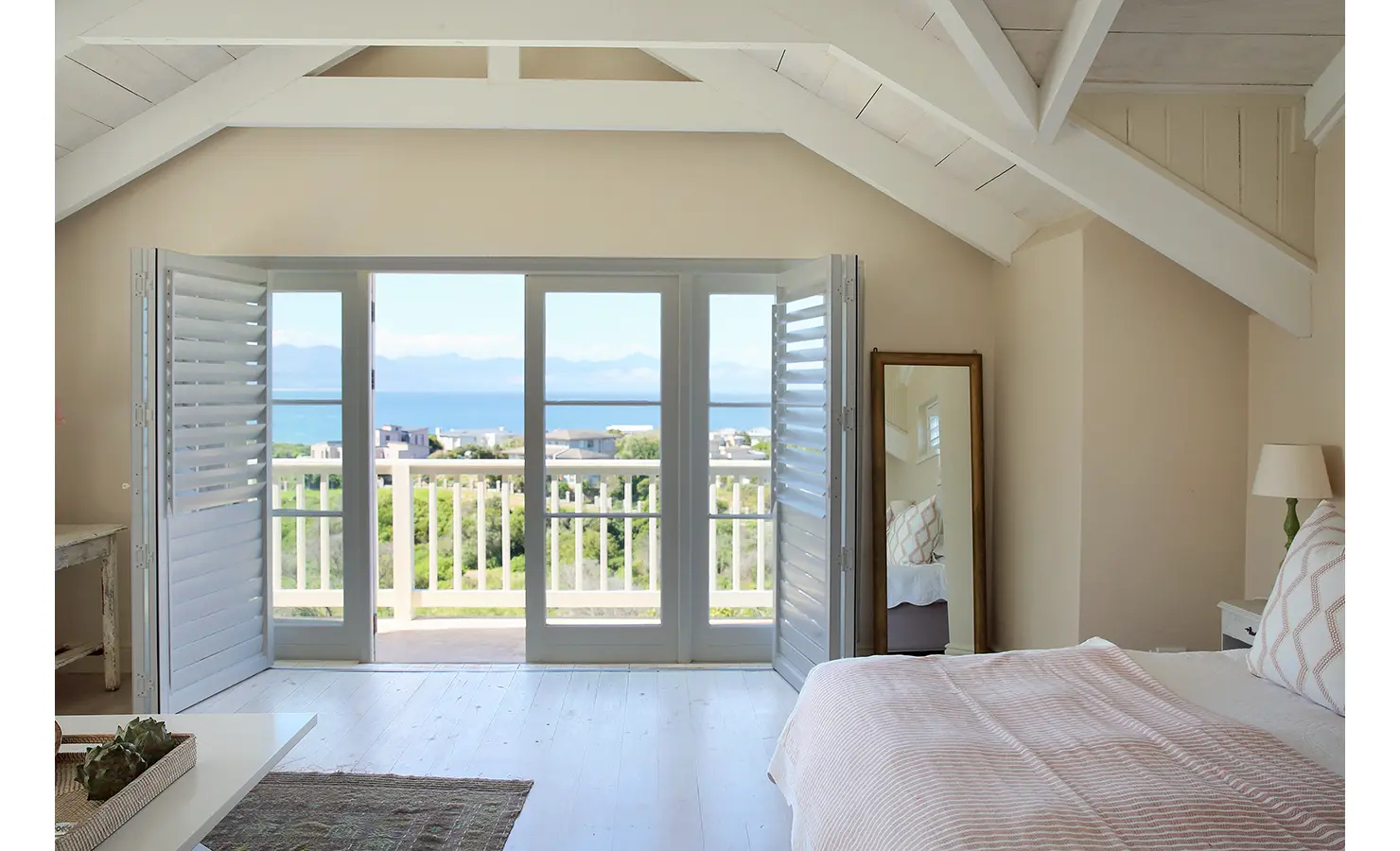 Perfect Hideaways: A bright bedroom with exposed beams and white shutters opening to a balcony that offers stunning ocean views. Beachwood On Robberg, Plettenberg Bay.