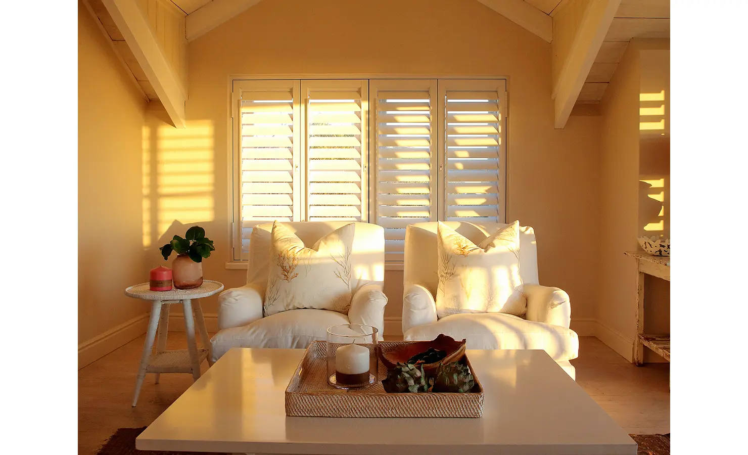 Perfect Hideaways: A tranquil sitting area bathed in golden sunlight, featuring white armchairs, decorative cushions, and plantation shutters. Beachwood On Robberg, Plettenberg Bay.