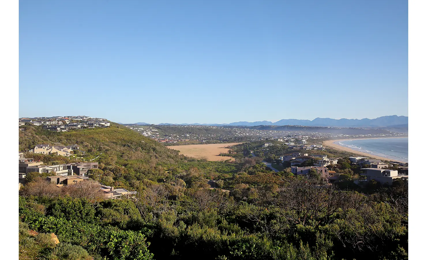 Perfect Hideaways: A panoramic view of the coastline, showcasing rolling hills, sandy beaches, and scattered luxury homes under a bright blue sky. Beachwood On Robberg, Plettenberg Bay.