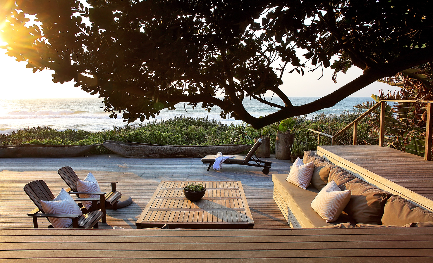 Perfect Hideaways: A serene wooden deck overlooking the ocean, shaded by a sprawling tree. The inviting outdoor seating area features plush cushions and Adirondack chairs, complemented by a sun lounger positioned for the perfect sunset view. Lush greenery frames the setting, enhancing the coastal charm. Cowrie Cottage, Westbrook Beach.