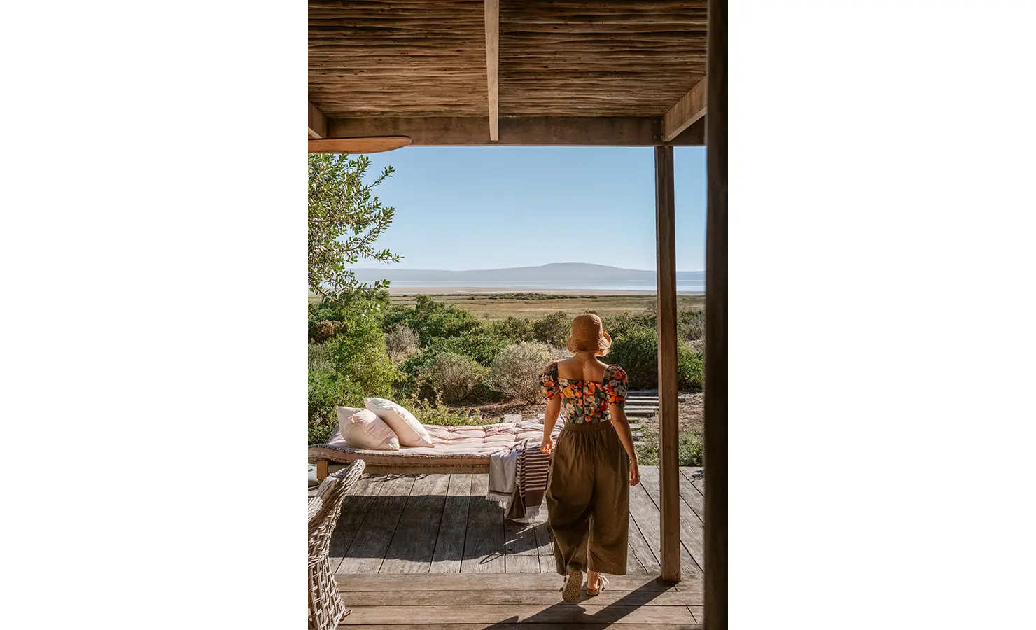 Perfect Hideaways: A woman in a straw hat and flowing skirt steps onto the deck, bathed in soft morning light, gazing across a fynbos-covered landscape towards the shimmering lagoon in the distance. A daybed layered with blush-toned cushions invites slow, sun-soaked moments. Yellowwood Cottage, Churchhaven.