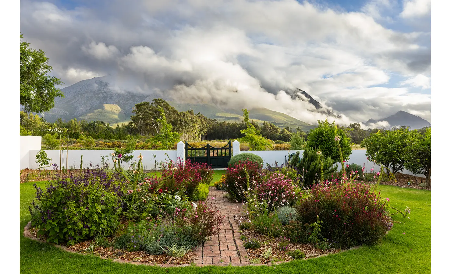 Perfect Hideaways: A lush garden in full bloom leads to a charming black gate, with misty mountains rising in the distance under dramatic cloud cover. A serene countryside retreat. Eikehof Farm, Worcester.
