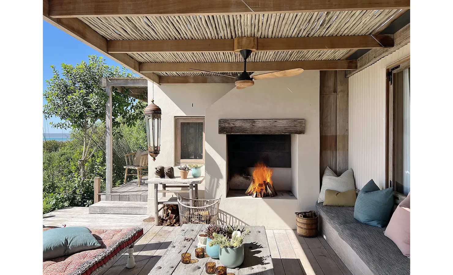 Perfect Hideaways: A rustic covered veranda with a crackling fire in the built-in hearth, framed by wicker chairs, plump scatter cushions and soft neutral tones. A daybed and potted succulents add charm to the relaxed outdoor living space. Yellowwood Cottage, Churchhaven.