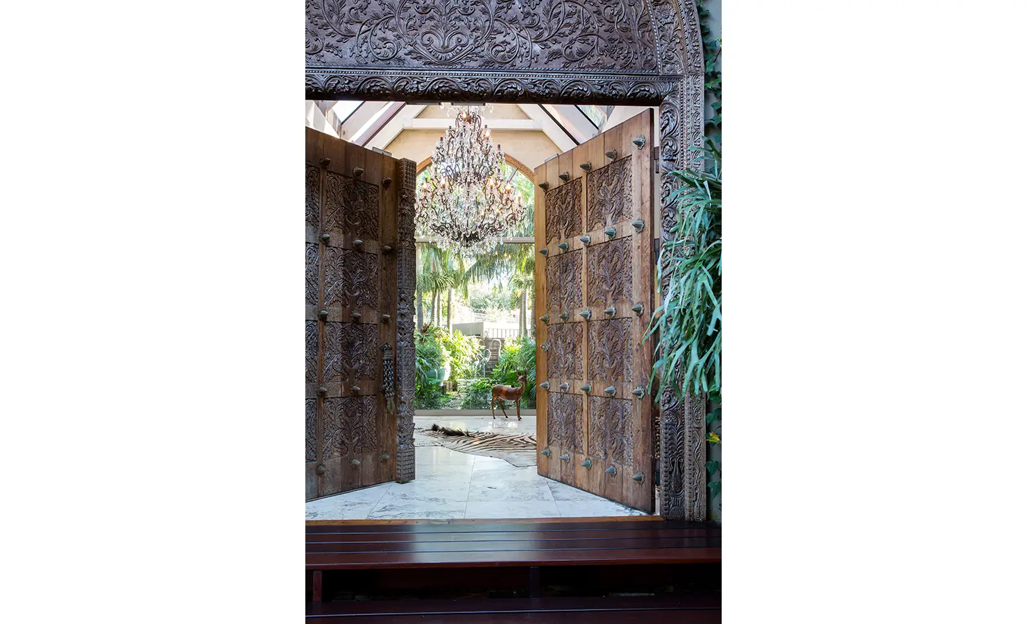 Perfect Hideaways: An intricately carved wooden doorway opens to a lush atrium, framed by trailing greenery. A grand chandelier hangs overhead, casting light onto the marble floors, creating an enchanting entrance into this tropical oasis. Sophias Retreat, Durban North.