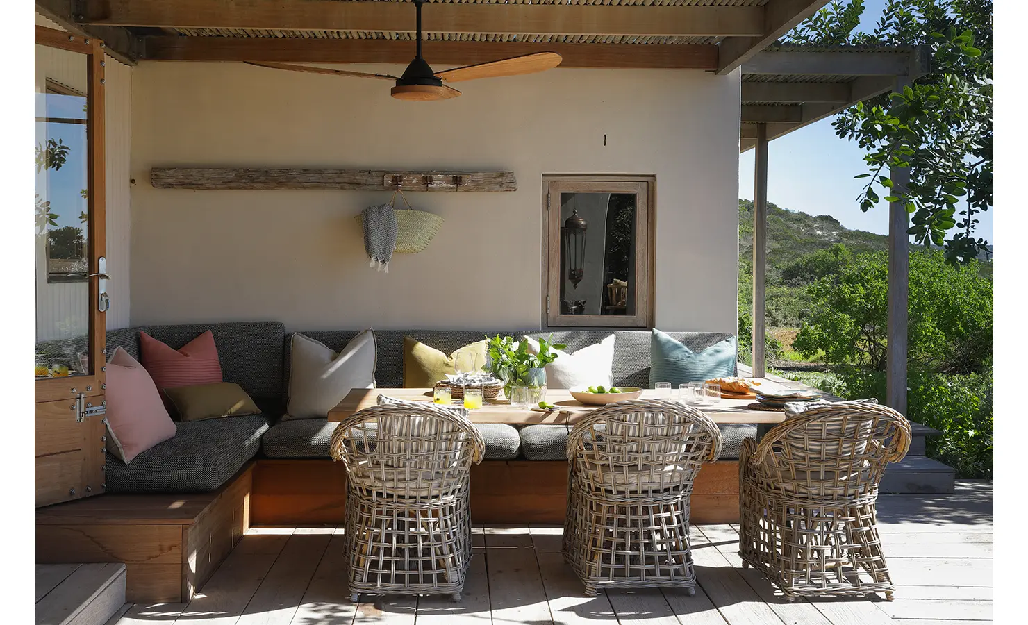 Perfect Hideaways: A cosy outdoor seating area featuring a wooden table, wicker chairs, and cushioned benches with colourful pillows, set under a shaded patio with greenery in the background. Yellowwood Cottage, Churchhaven.