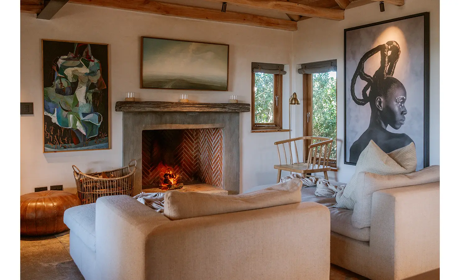 Perfect Hideaways: An inviting living room with a roaring fireplace and textured herringbone brickwork, flanked by modern African portraiture and abstract artworks. Neutral-toned sofas and a basket of firewood complete the cocooning ambience. Yellowwood Cottage, Churchhaven.