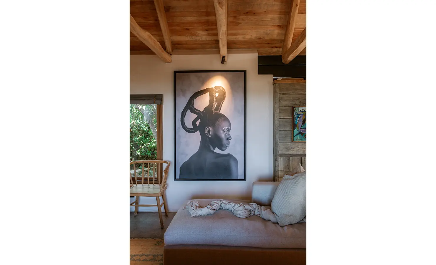Perfect Hideaways: A close-up view of a large monochrome portrait of a woman with sculptural hair, elegantly lit above a daybed, surrounded by raw timber walls and windows that frame the surrounding greenery. Yellowwood Cottage, Churchhaven.