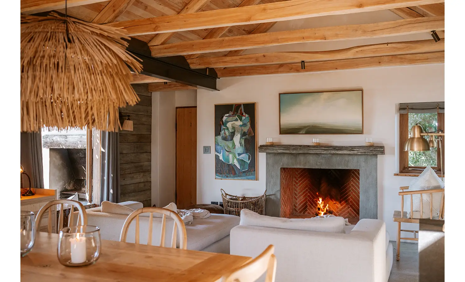 Perfect Hideaways: A warm, open-plan living space anchored by a roaring fire and soft cream sofas, accented by curated artworks and layered textures. Natural wood tones and woven lighting infuse the room with relaxed sophistication. Yellowwood Cottage, Churchhaven.