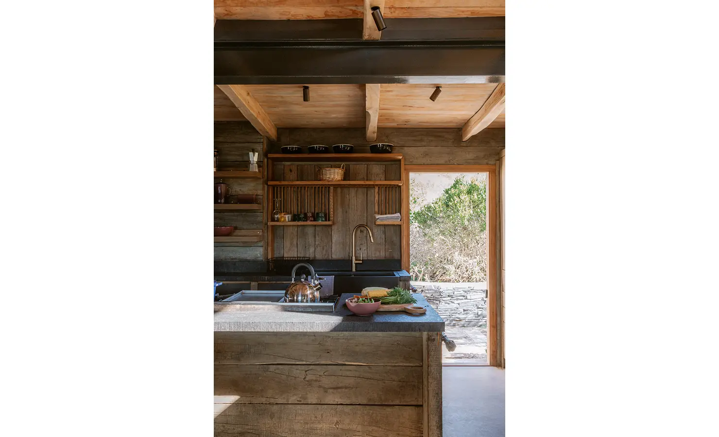 Perfect Hideaways: A moody yet elegant kitchen with open wooden shelving, raw timber cabinetry and stone countertops, opening directly to a path beyond. A bowl of produce and steaming kettle evoke a lived-in, homely feel. Yellowwood Cottage, Churchhaven.
