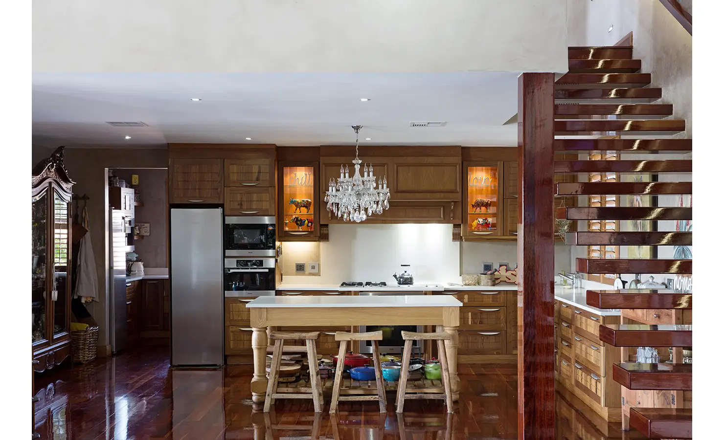 Perfect Hideaways: A warm and inviting kitchen with rich wooden cabinetry, a central island with rustic stools, and a sparkling crystal chandelier. A sleek floating staircase with glossy wood steps leads to the upper level, enhancing the modern-rustic aesthetic. Sophias Retreat, Durban North.