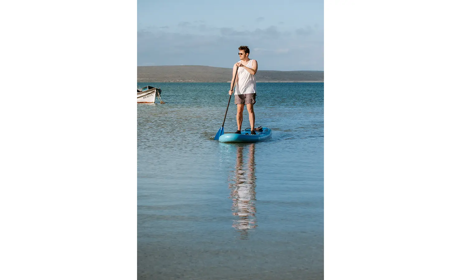 Perfect Hideaways: A man enjoys the calm waters of the lagoon while paddleboarding on a bright blue board, surrounded by nature and quiet solitude. A small fishing boat drifts nearby, reinforcing the peaceful coastal lifestyle. Yellowwood Cottage, Churchhaven.
