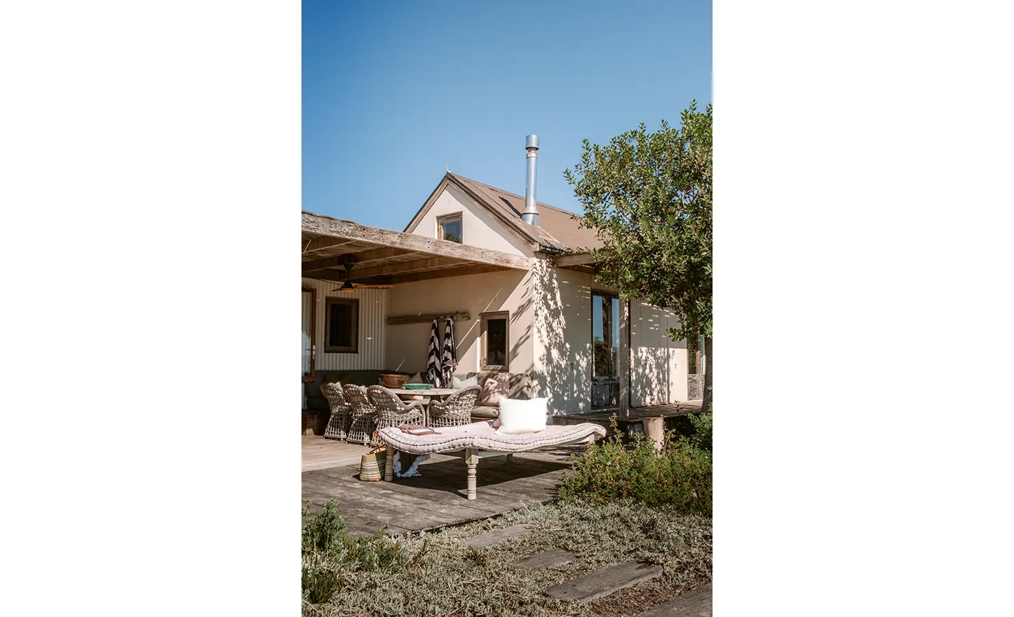 Perfect Hideaways: The sun-drenched exterior of the cottage features a shaded veranda with a dining table, wicker chairs and a cushioned daybed, framed by indigenous flora and the wide blue sky above. Yellowwood Cottage, Churchhaven.