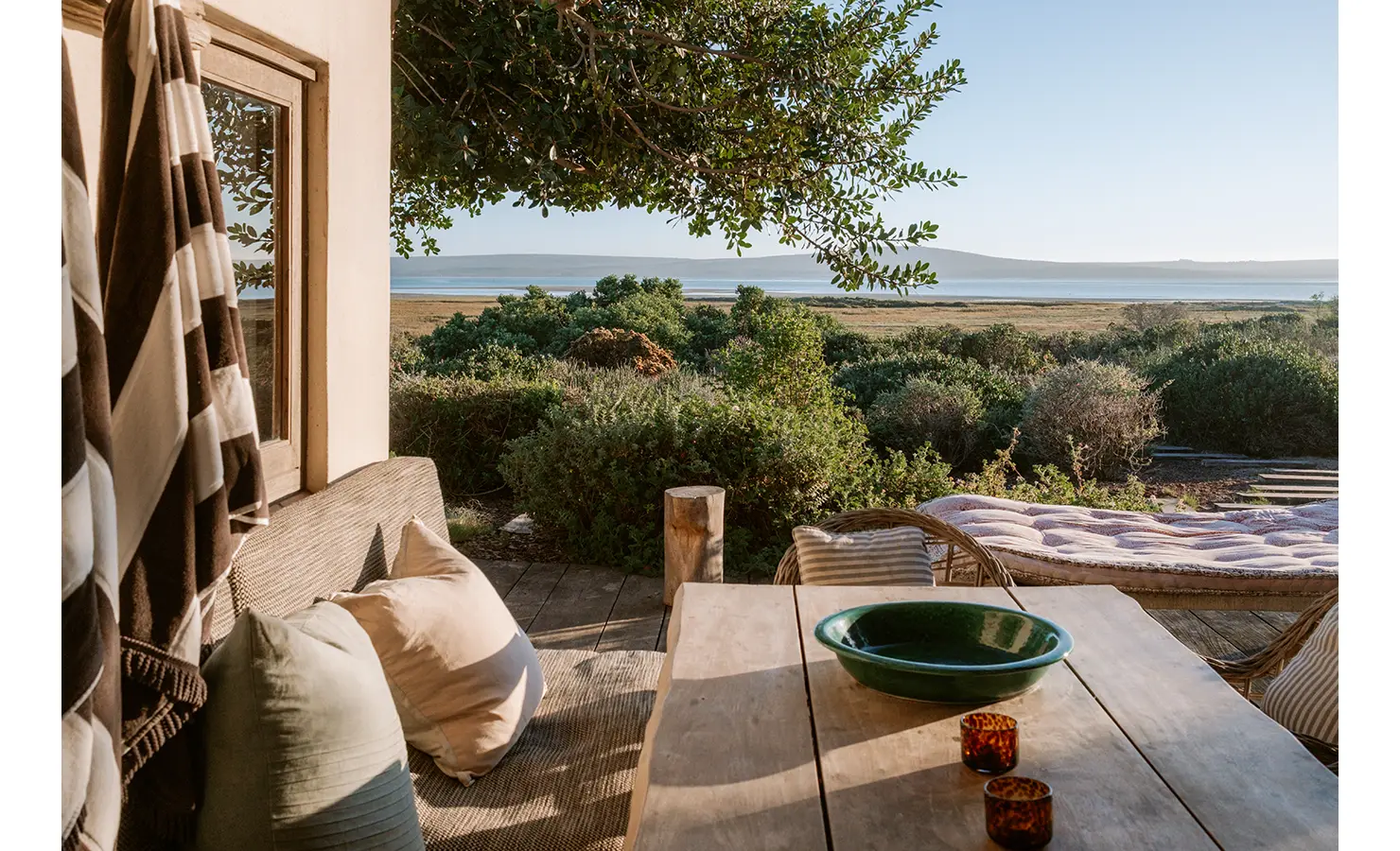 Perfect Hideaways: A peaceful veranda scene at golden hour, with sunlight casting warm shadows over a wooden dining table, striped beach towel, and tufted daybed, all overlooking a wild fynbos garden and still lagoon. Yellowwood Cottage, Churchhaven.