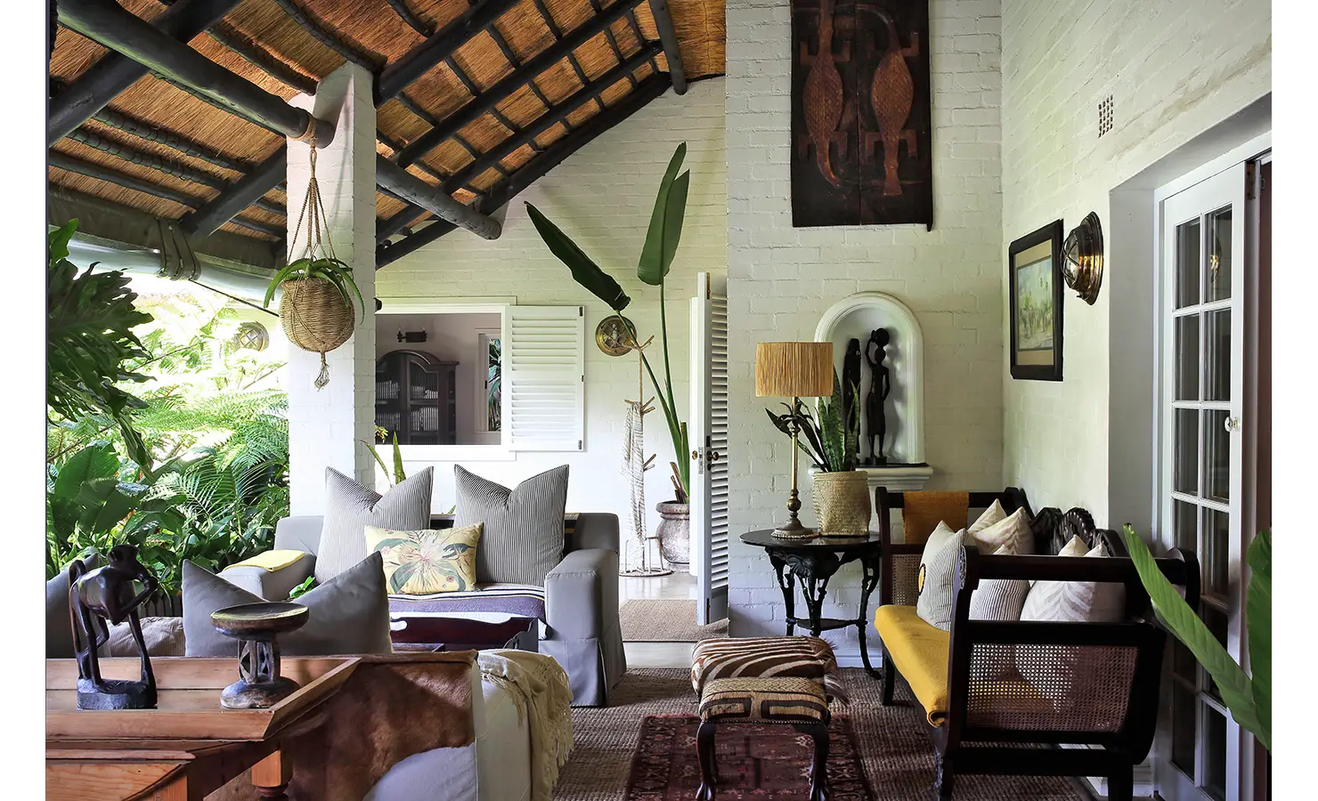 Perfect Hideaways: A beautifully styled open-plan living space under a thatched roof, blending natural textures with curated African décor. Woven furniture, soft cushions, and lush greenery create an inviting, tranquil retreat. Cowrie Cottage, Westbrook Beach.