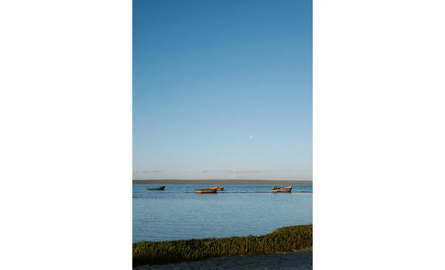 Perfect Hideaways: Traditional wooden fishing boats float gently on the still lagoon beneath a soft morning sky, the pale moon still visible above the distant horizon. Yellowwood Cottage, Churchhaven.
