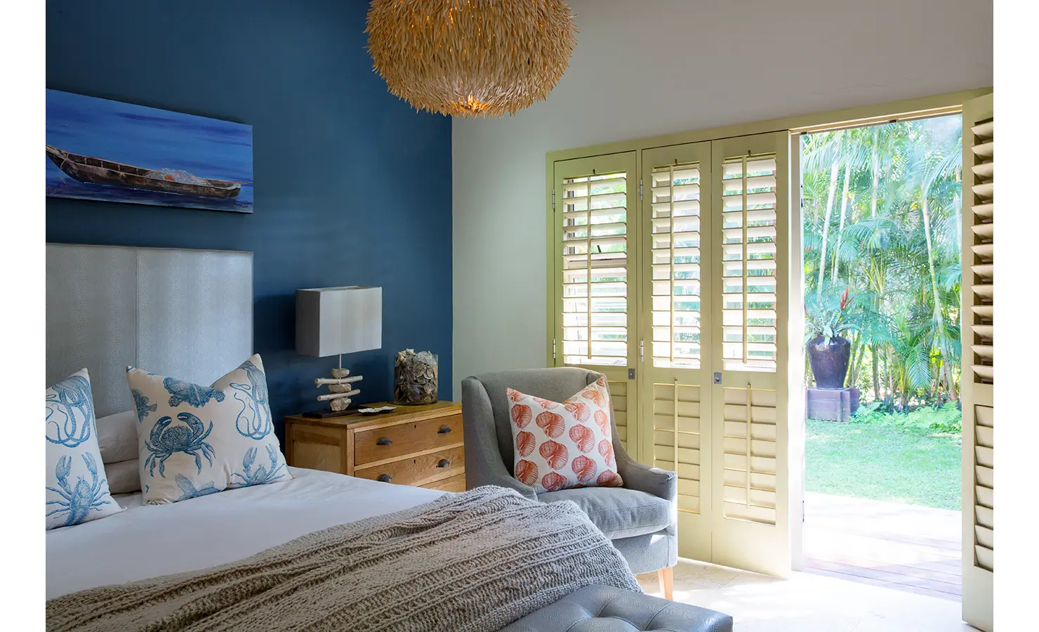 Perfect Hideaways: A tranquil bedroom with a soothing coastal theme, featuring a navy accent wall, sea-inspired decor, and a woven pendant light. Wooden shutters open onto a lush garden, bringing the outdoors into this serene space. Sophias Retreat, Durban North.