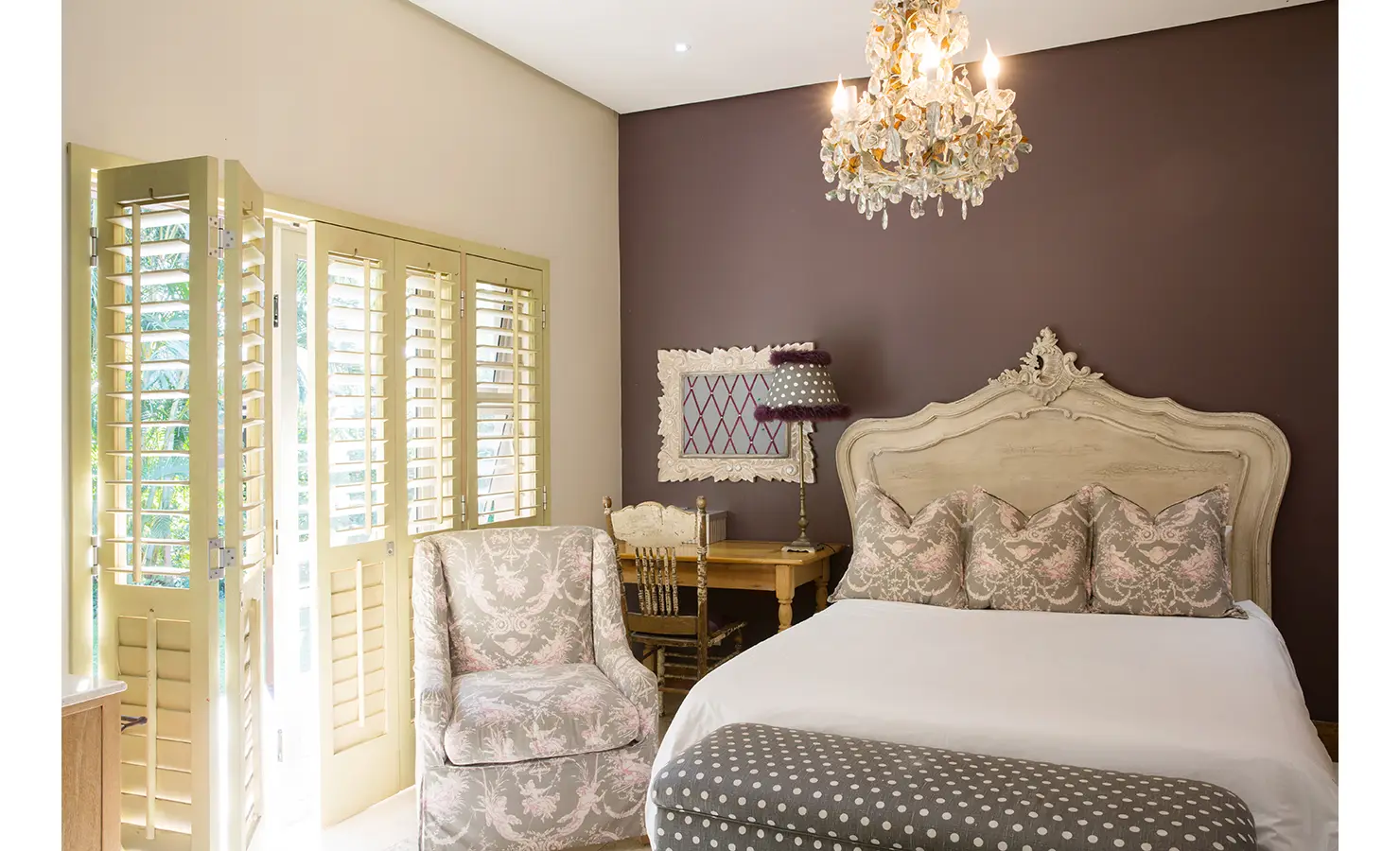 Perfect Hideaways: A charming bedroom with an antique-style wooden bed, soft floral-patterned cushions, and a delicate crystal chandelier. Light filters through the classic shutters, illuminating the warm, vintage-inspired decor. Sophias Retreat, Durban North.