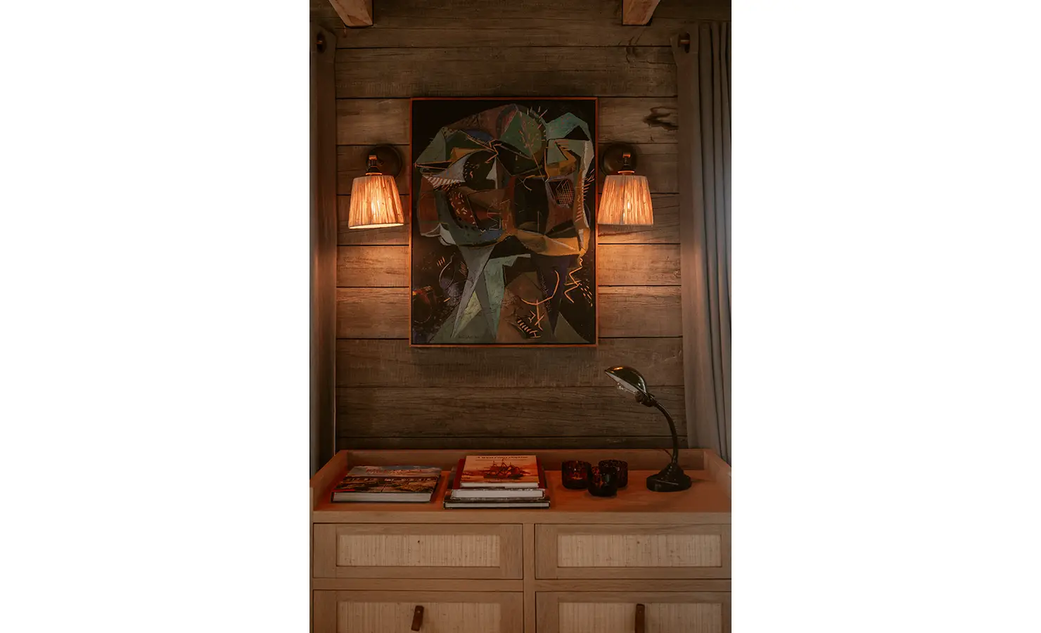 Perfect Hideaways: A cosy reading nook glows with warm lamplight against timber-panelled walls, with artful abstract portraiture and a curated collection of books and votives on a wooden dresser. Yellowwood Cottage, Churchhaven.