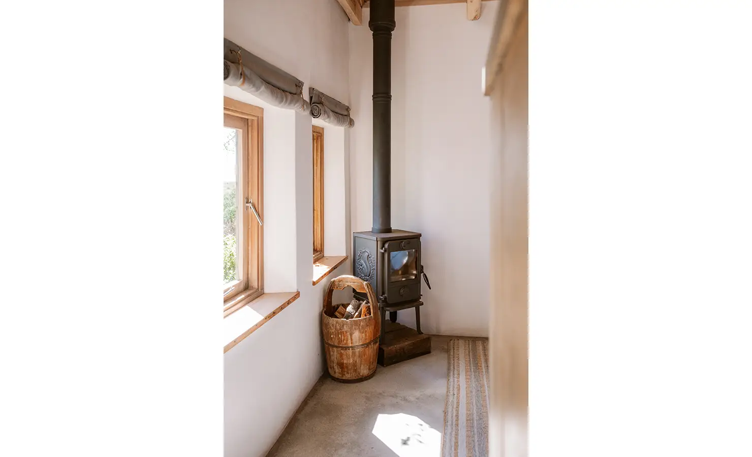 Perfect Hideaways: A vintage cast-iron wood-burning stove is tucked into the corner of a whitewashed room, surrounded by hand-plastered walls, warm sunlight, and a basket of neatly stacked firewood. Yellowwood Cottage, Churchhaven.