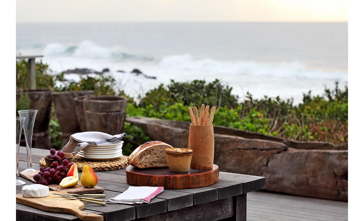 Perfect Hideaways: A beautifully styled outdoor dining table set with fresh fruit, rustic bread, and crisp white plates, overlooking the rolling ocean waves. The perfect setting for a leisurely seaside meal. Cowrie Cottage, Westbrook Beach.