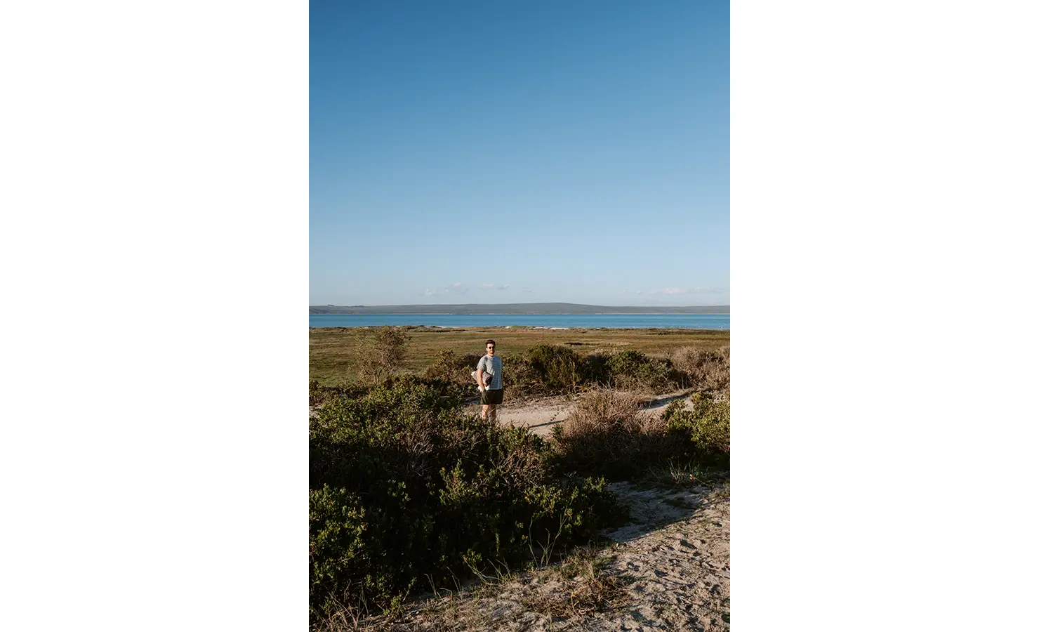 Perfect Hideaways: A man walks a sandy path through fynbos down towards the water’s edge, the expansive lagoon and distant hills stretching beneath a vast, cloudless sky. Yellowwood Cottage, Churchhaven.