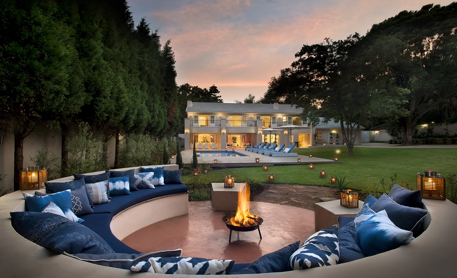 Perfect Hideaways: A firepit lounge with indigo cushions creates a cosy evening ambiance in the landscaped garden of a grand, double-storey villa with glowing lights, a pool terrace, and elegant symmetry at sunset. AtholPlace Hotel & Villa, Atholl.