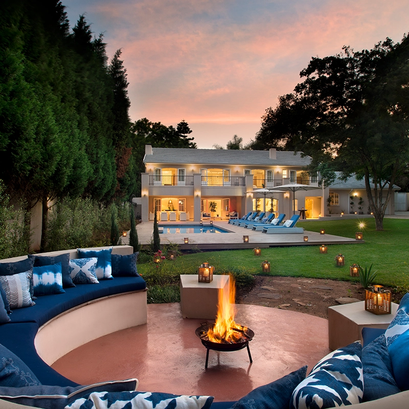 Perfect Hideaways: A firepit lounge with indigo cushions creates a cosy evening ambiance in the landscaped garden of a grand, double-storey villa with glowing lights, a pool terrace, and elegant symmetry at sunset. AtholPlace Hotel & Villa, Atholl.