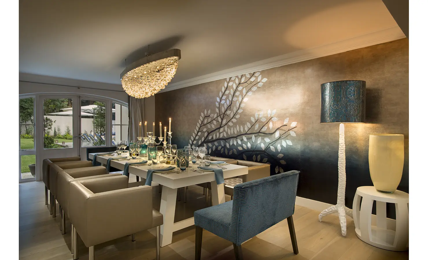 Perfect Hideaways: A sophisticated dining room features a striking crystal chandelier, a wall mural of metallic leaves, and a candlelit table set for a glamorous dinner party. AtholPlace Hotel & Villa, Atholl.