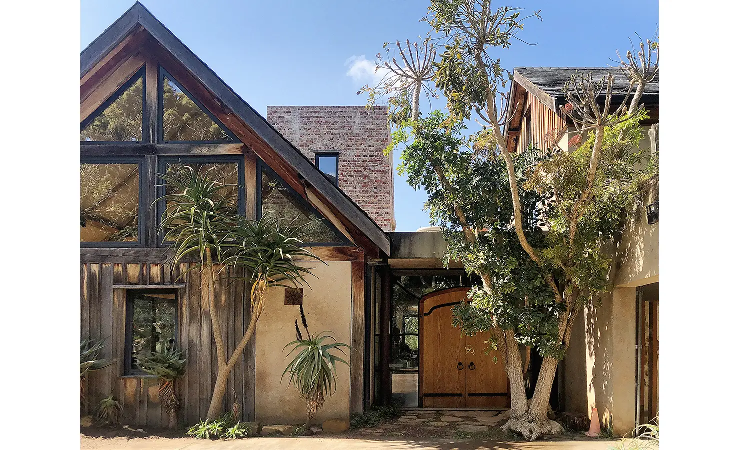 Perfect Hideaways. Exterior view of a rustic modern house with large windows and wooden accents, surrounded by lush greenery and trees against a clear blue sky. Gaia, Constantia.